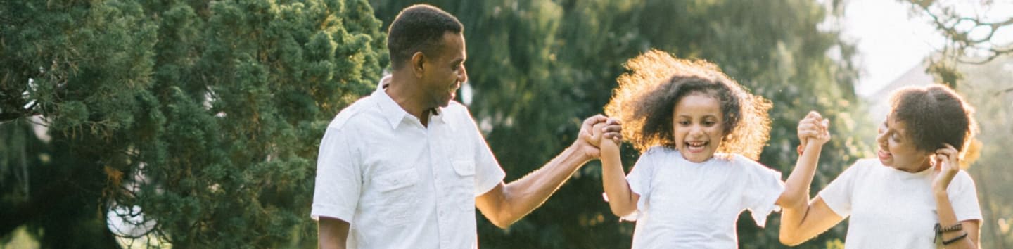A smiling family wearing white clothes holds hands in a lush, green park.