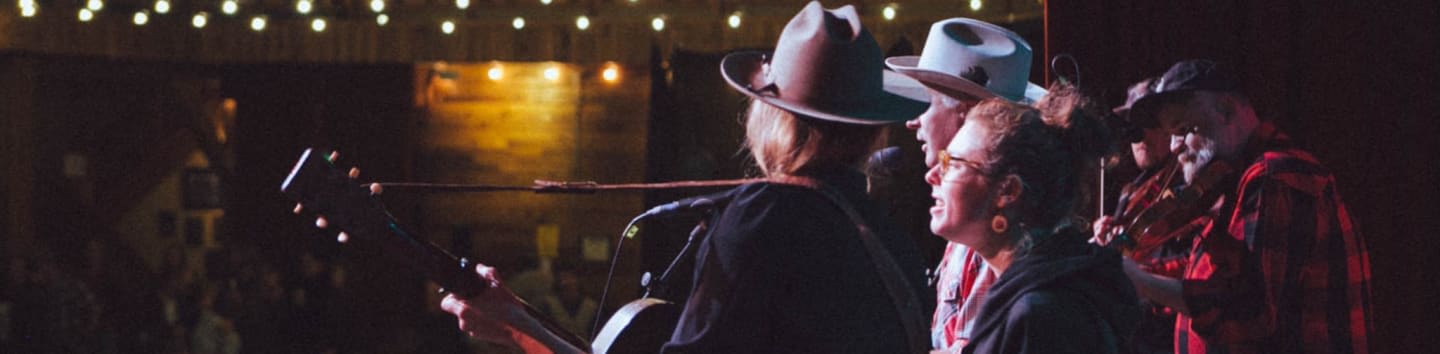 A group of musicians wearing cowboy hats performs on a dimly lit stage against a background of warm string lights and wooden walls.