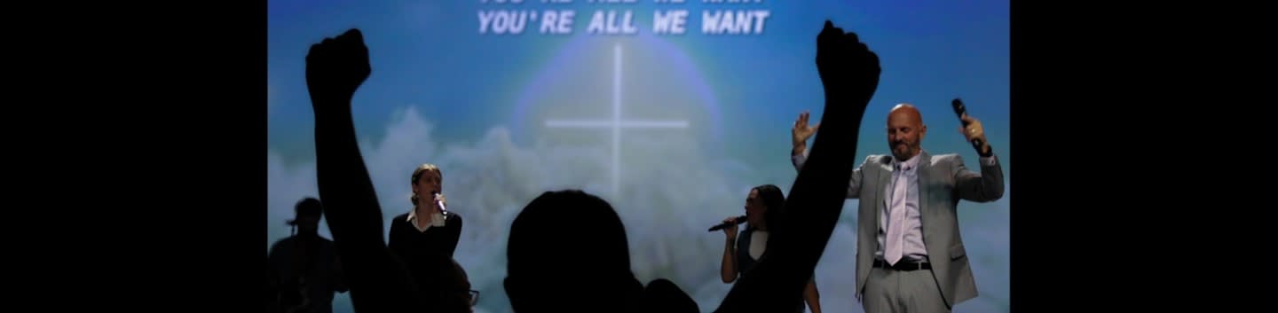 Silhouetted arms are raised in the foreground before a stage where singers perform against a background screen featuring a cross and the text "YOU'RE ALL WE WANT."