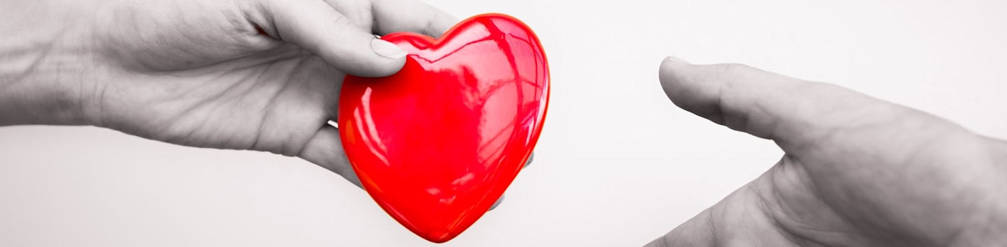 A grayscale hand holds a vibrant red heart toward another grayscale hand against a plain white background.