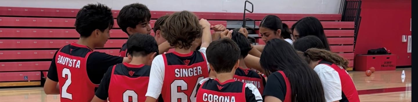 A group of young athletes in red and black uniforms huddle together in the foreground against a background of red bleachers.