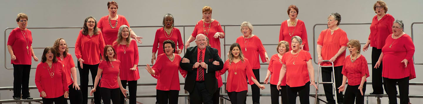 A choir of women in red tops and a man in a black suit stand and sing against a plain white background.
