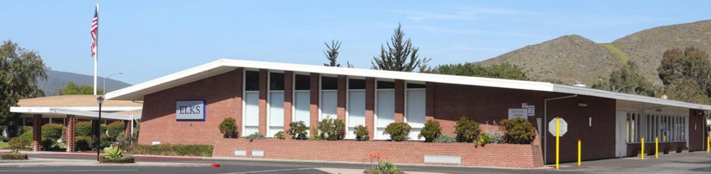 A brick Elks lodge building sits in the foreground under a clear blue sky, with rolling hills and trees in the background.