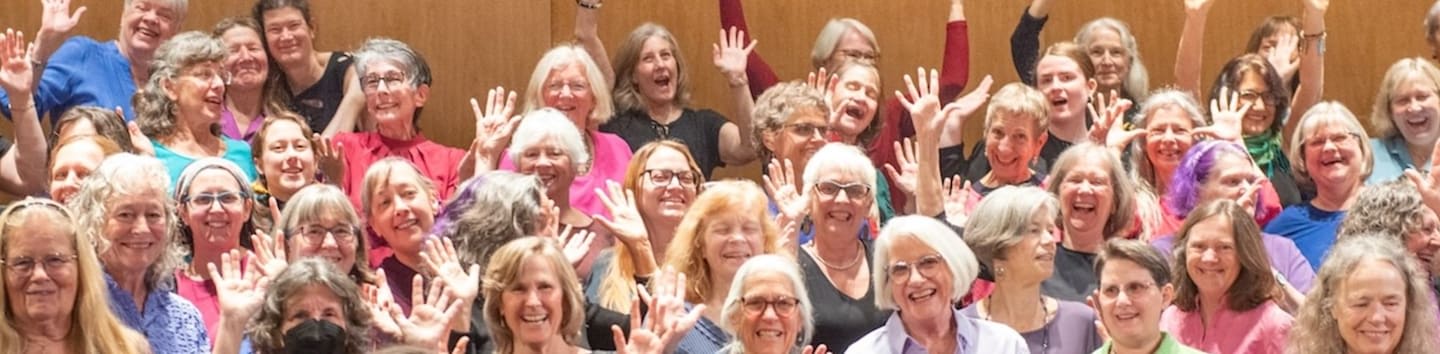 A large group of smiling women with their hands raised fills the foreground against a plain, light-brown wall in the background.