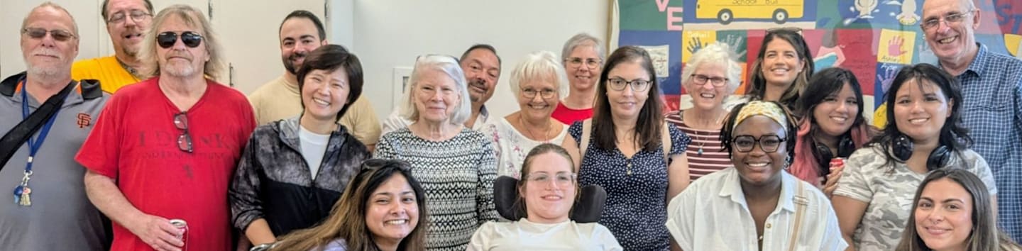 A diverse group of people smile for a photo in the foreground against a plain white wall and a colorful children's mural in the background.
