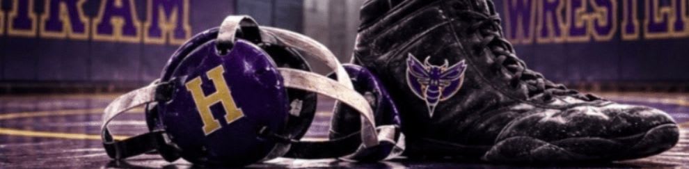 A wrestling shoe and headgear rest on a mat in front of a blurred background of a wrestling arena with the Hiram Hornets logo.