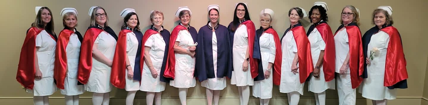 A group of women dressed in white nurse uniforms and red and blue capes stand in a line against a plain, neutral-colored wall.