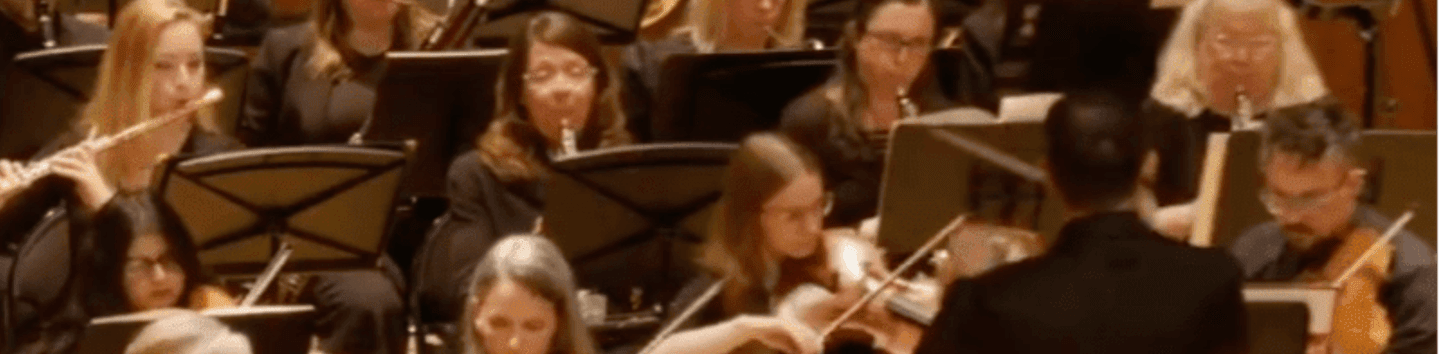 An orchestra of musicians playing string and brass instruments performs under the direction of a conductor in a wood-paneled concert hall.