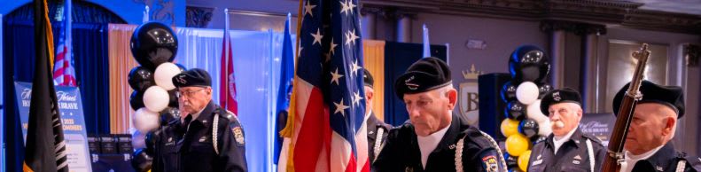 In the foreground, several men in uniform stand with an American flag, while the background features a decorated event hall with balloons and banners.