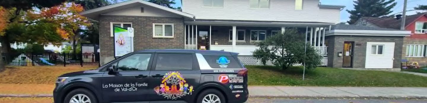 A dark blue branded minivan is parked on a street in front of a white and grey two-story house with a green lawn.