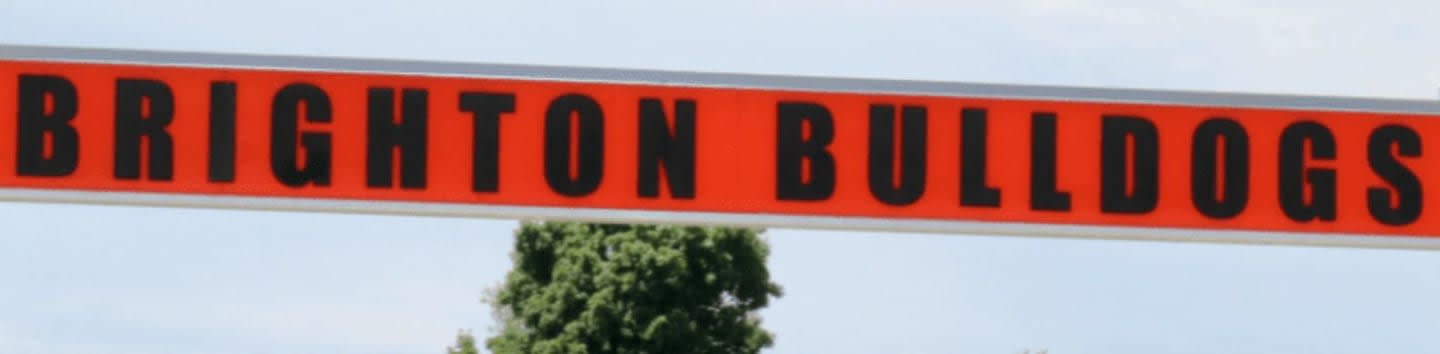 An orange and black "BRIGHTON BULLDOGS" sign hangs above a black metal fence with green trees and a white building in the background.