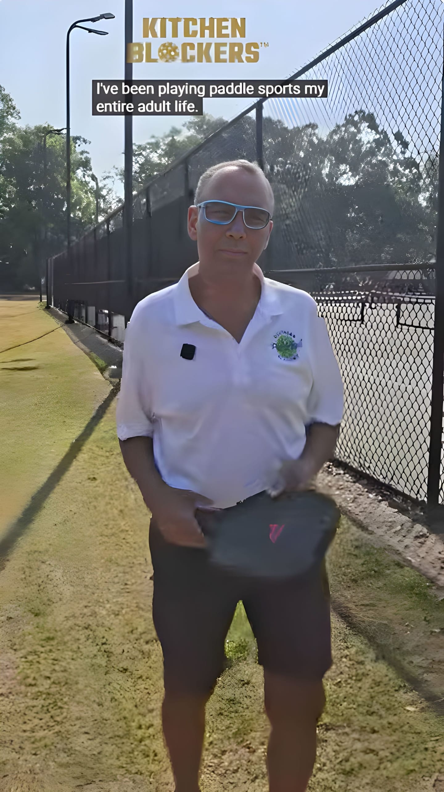 A man in a white polo shirt and sunglasses stands on a pickleball court holding a paddle, with a fence and trees in the background.