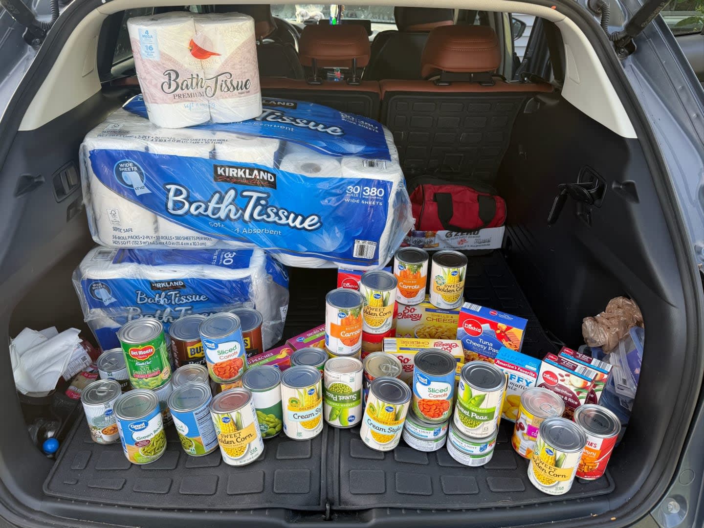 A car trunk is filled with a large supply of toilet paper and canned goods, with a red first-aid kit visible in the background.