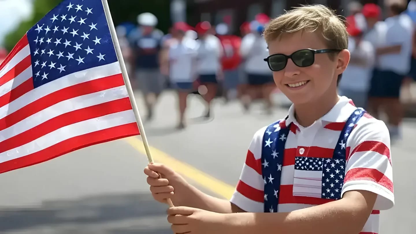 A smiling boy in patriotic attire holds an American flag in the foreground, while a blurred parade of people marches in the background.