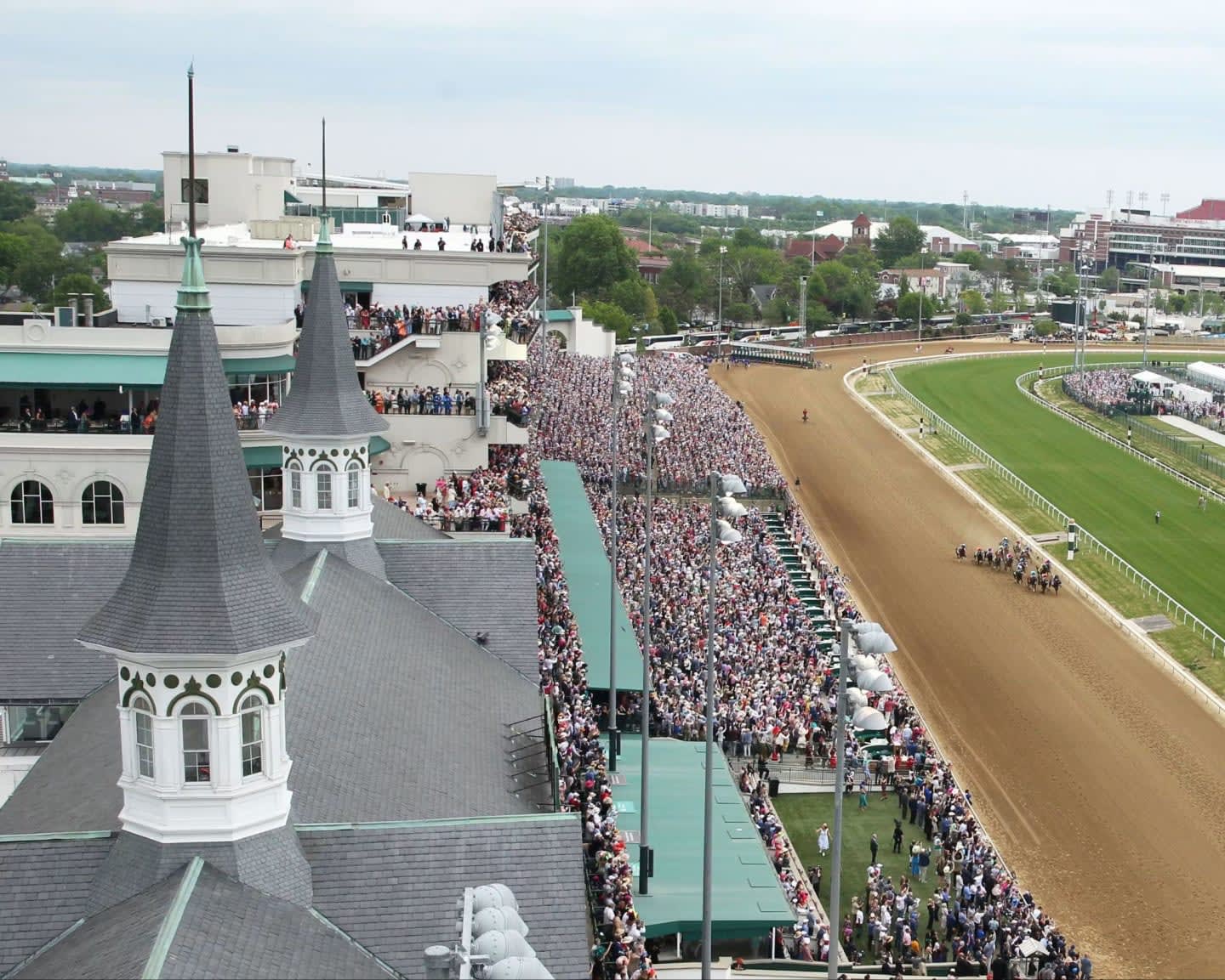 A horse race is underway on a dirt track with a large crowd gathered in the grandstands, while distinctive spired buildings dominate the foreground.