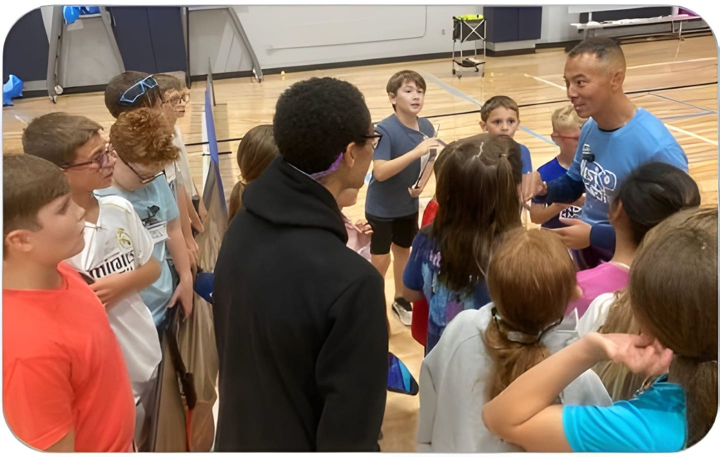 A group of children in the foreground are gathered around a man in a blue shirt in a gymnasium, with a tennis net and equipment visible in the background.