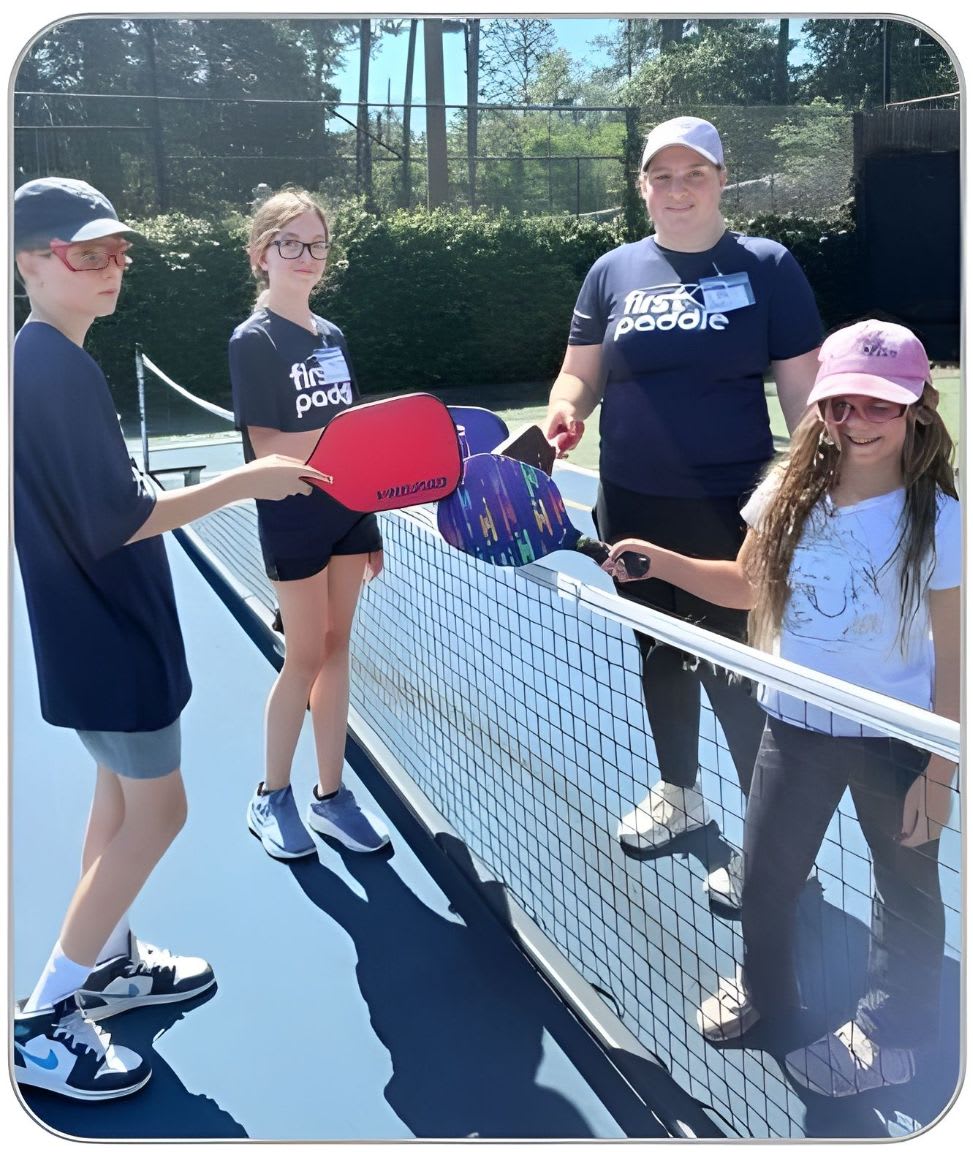 Four children and an adult stand on a blue pickleball court with paddles in hand, facing each other over a net, with trees and a fence in the background.