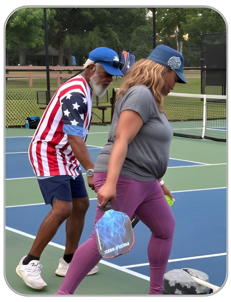 A man in a patriotic shirt and a woman in athletic wear walk across a pickleball court with a net and fence in the background.