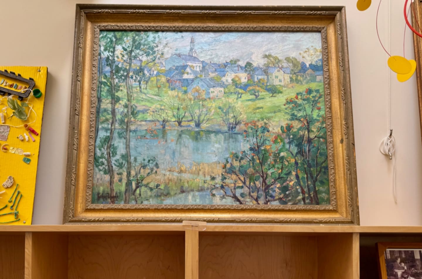 A framed impressionistic painting of a village by a lake is displayed on a shelf, with a yellow craft project to its left and a white wall behind.