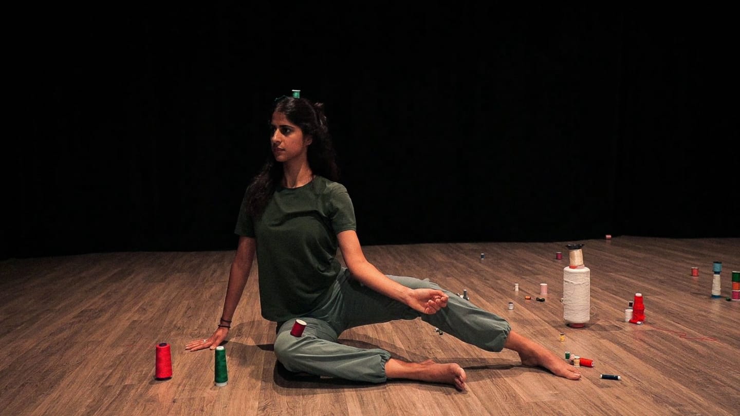 A woman in green attire sits on a wooden stage surrounded by spools of thread, with a dark backdrop behind her.