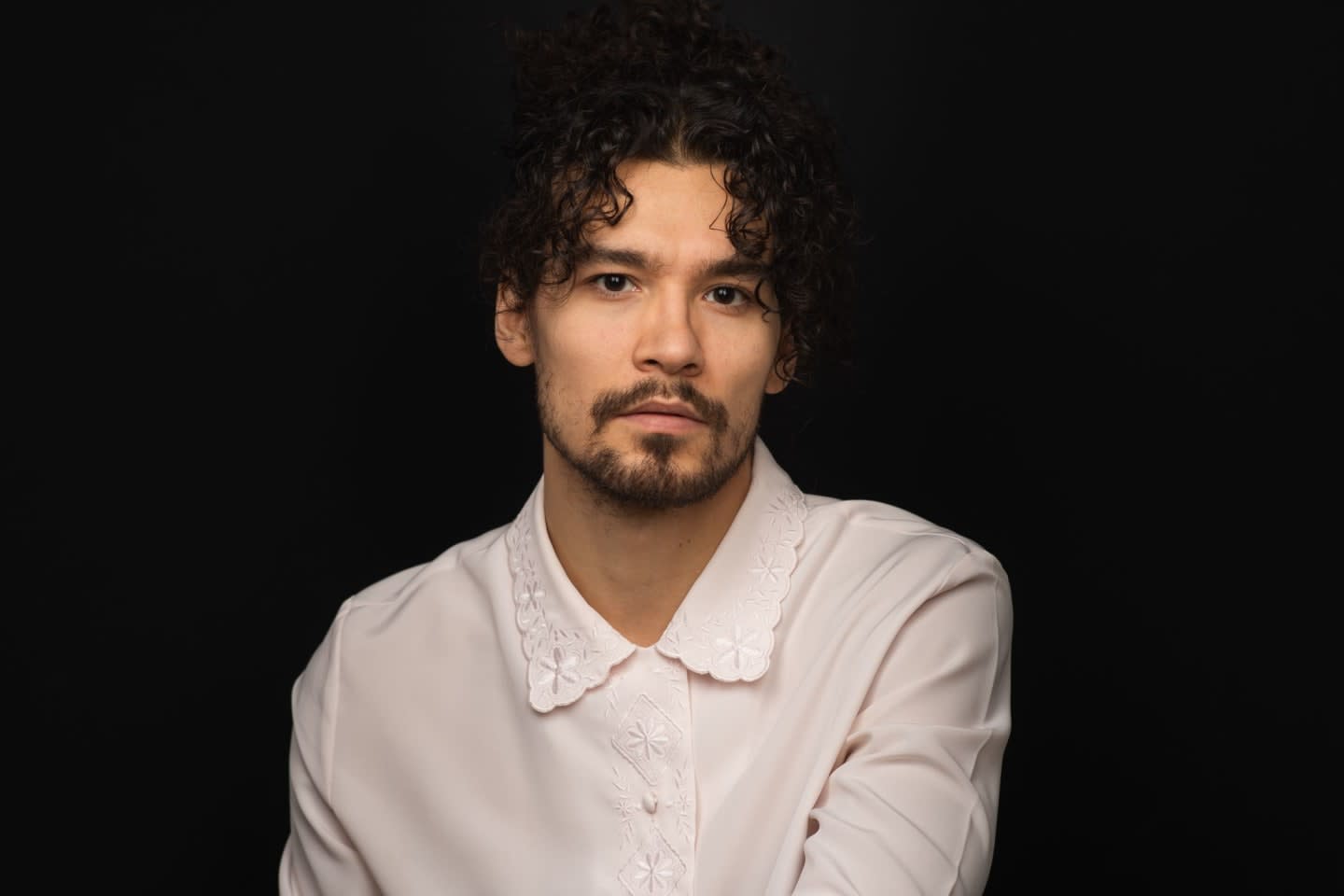 A man with curly dark hair and a beard, wearing a light-colored shirt with a decorative collar, is pictured against a plain black background.
