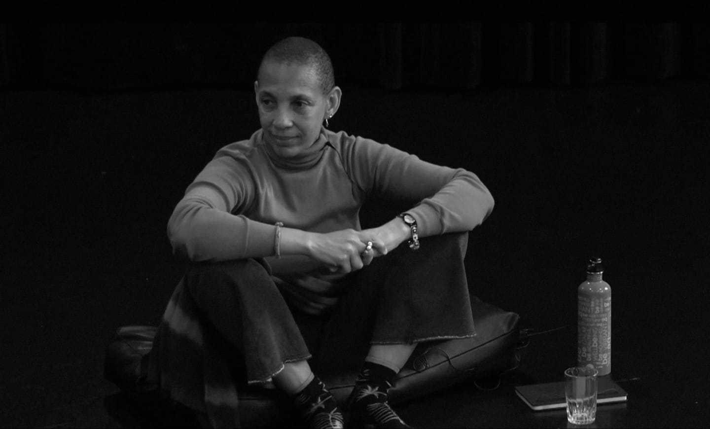 A bald woman in a turtleneck and patterned socks sits cross-legged on a cushion in the foreground, with a bottle and glass to her right, against a dark, out-of-focus background.