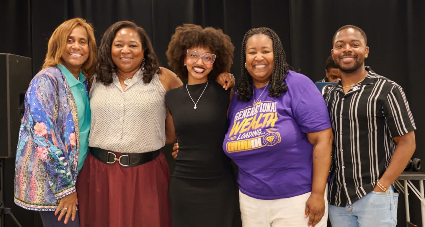 Five people stand smiling in front of a black curtain, with a man in a striped shirt on the right and a woman in a purple shirt with "GENERATIONAL WEALTH LOADING" on it to his left.