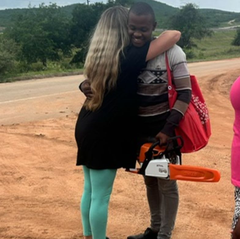A woman with long blonde hair hugs a young man holding a chainsaw, with a dirt road and trees in the background.