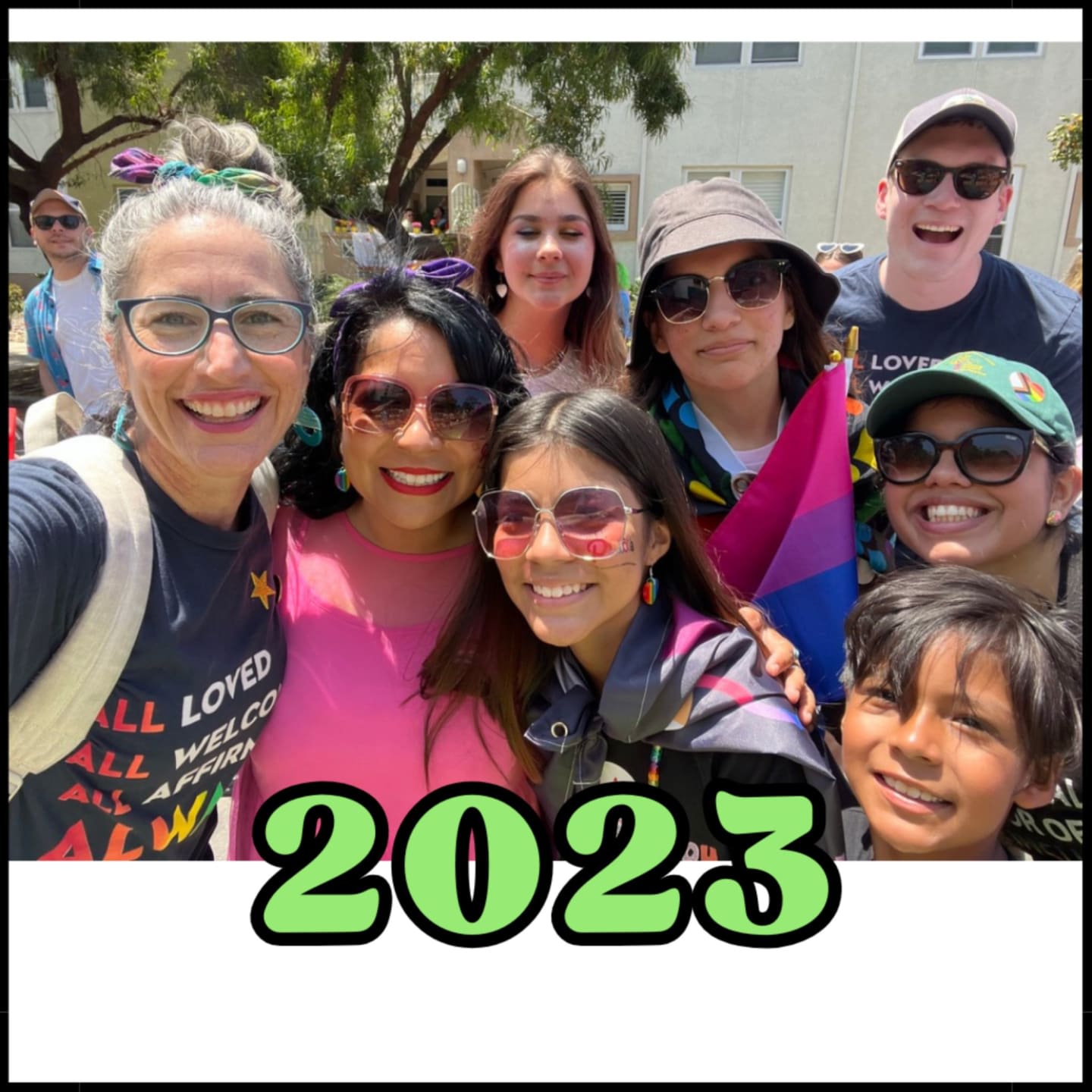 A group of smiling people, some wearing sunglasses and hats, pose for a selfie in front of a building with trees and a white border.