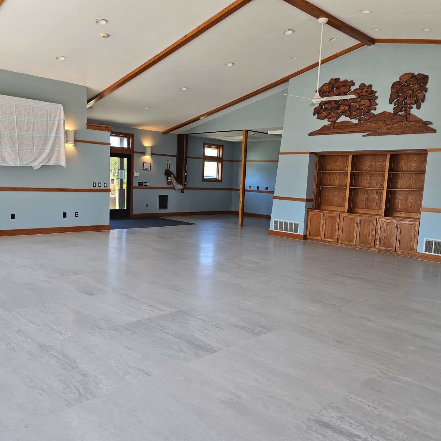 A large, empty room with a light gray floor in the foreground and light blue walls with wooden beams and a carved tree mural in the background.