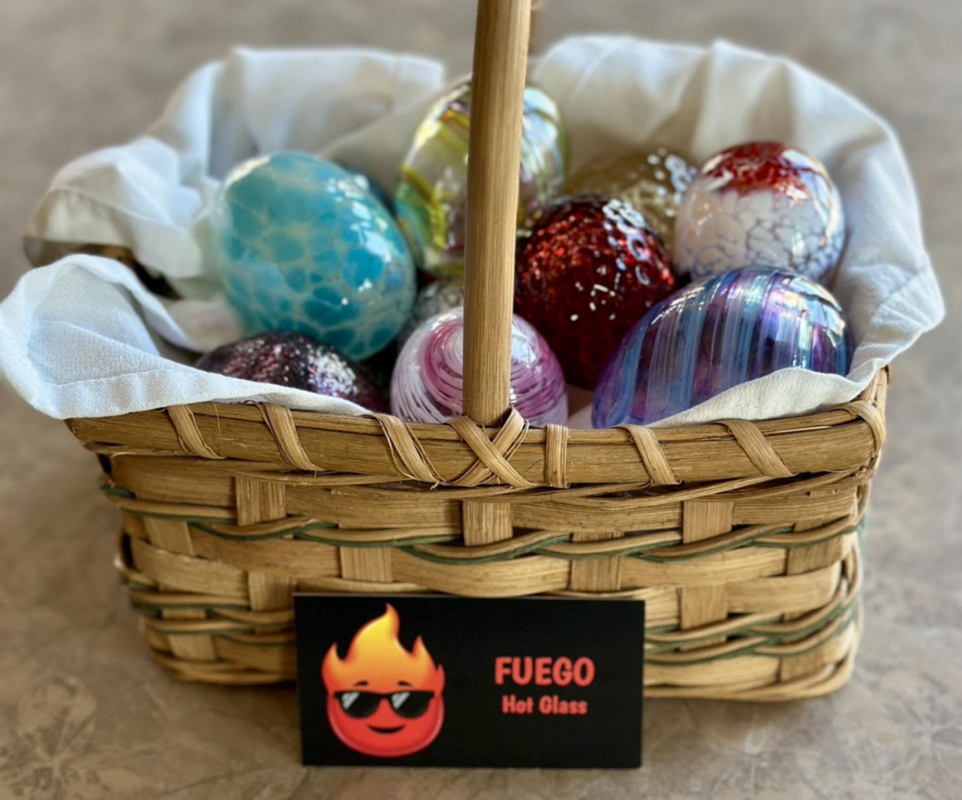 A woven basket filled with colorful, blown-glass eggs sits on a textured surface, with a "Fuego Hot Glass" business card in the foreground.