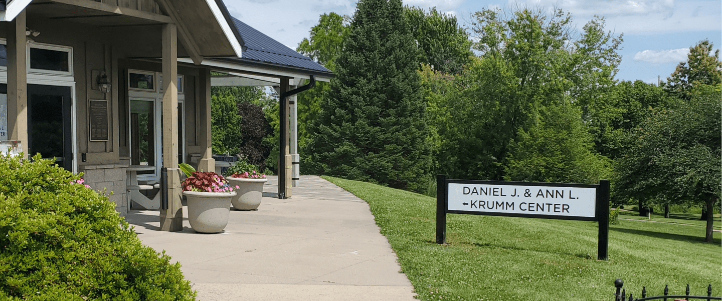 A paved walkway leads past a building with planters in the foreground, towards a sign for the Daniel J. & Ann L. Krumm Center surrounded by lush green trees in the background.