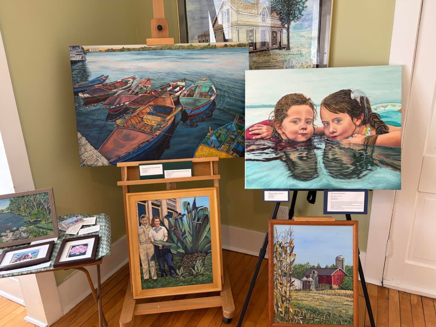 Several paintings of boats on water, two girls in a pool, a family portrait, and a barn are displayed on easels and a table in a room with light green walls and wooden floors.
