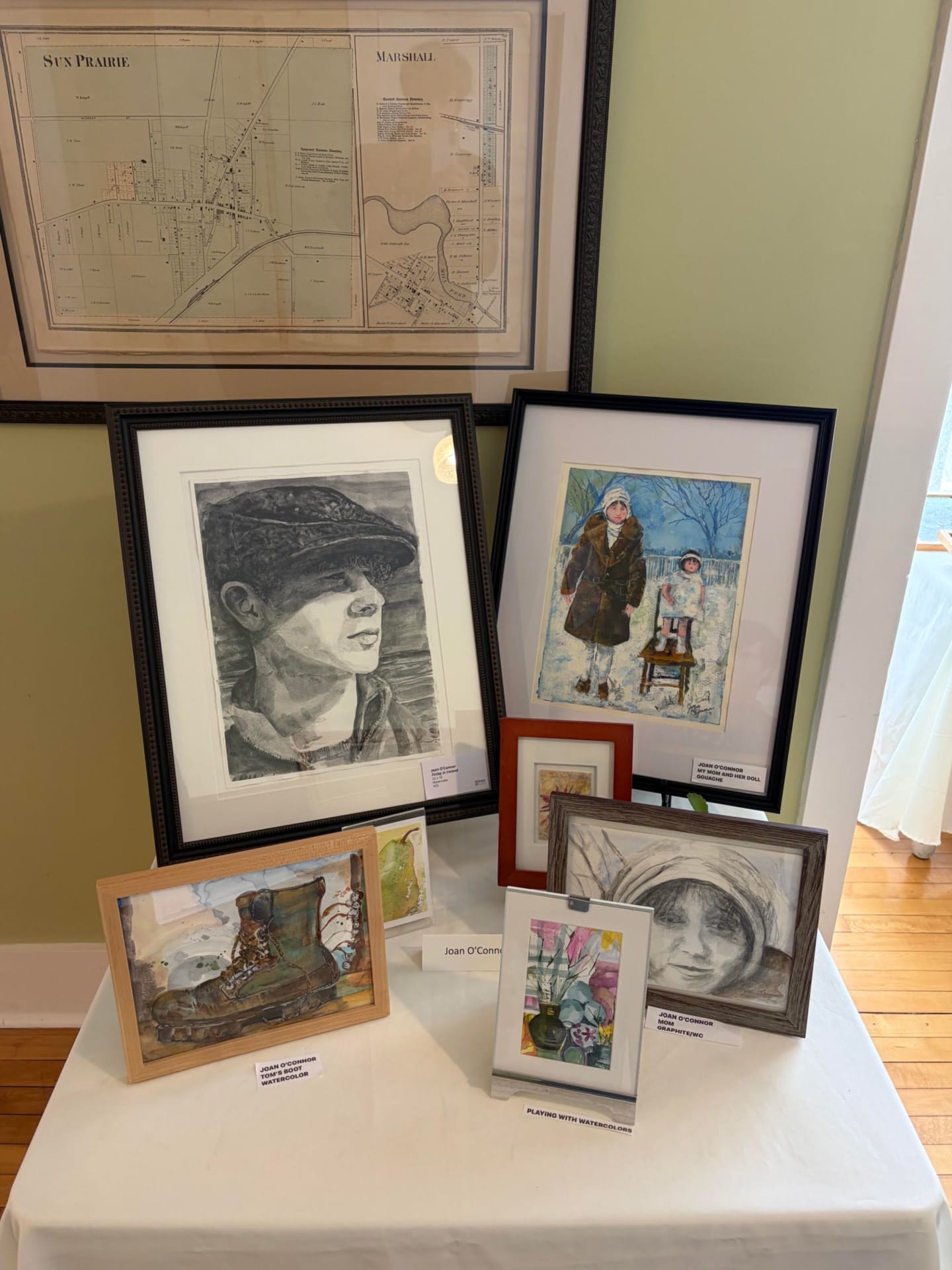 Several framed artworks, including portraits and a boot, are displayed on a white tablecloth in the foreground, with an antique map hanging on the wall in the background.