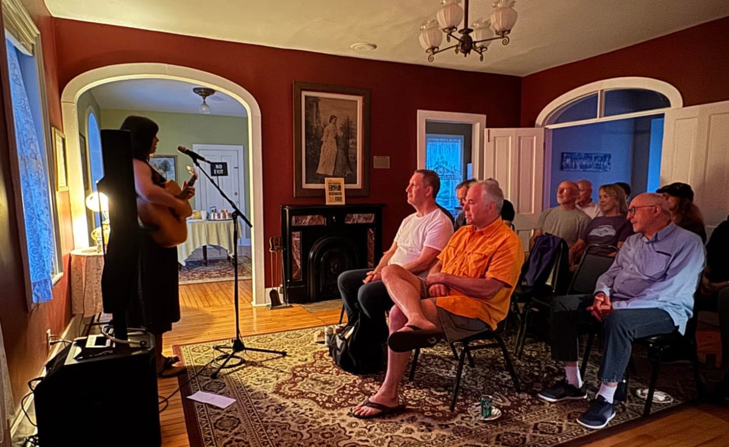 A woman plays guitar in the foreground while an audience watches in the background of a dimly lit room.