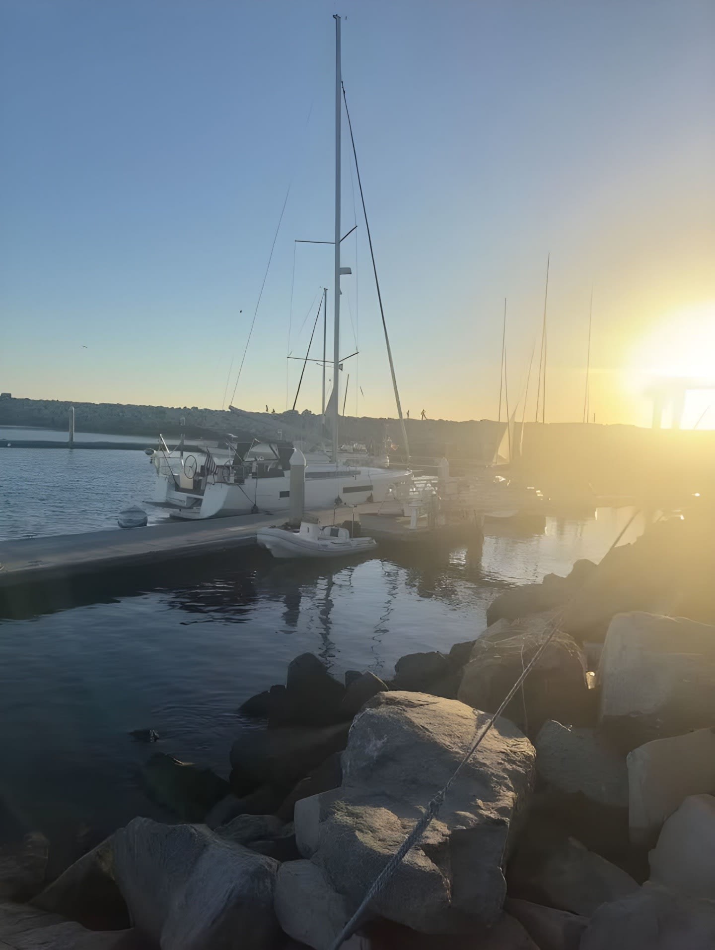 Large rocks line the foreground of a marina where sailboats are docked under a bright, hazy sun.