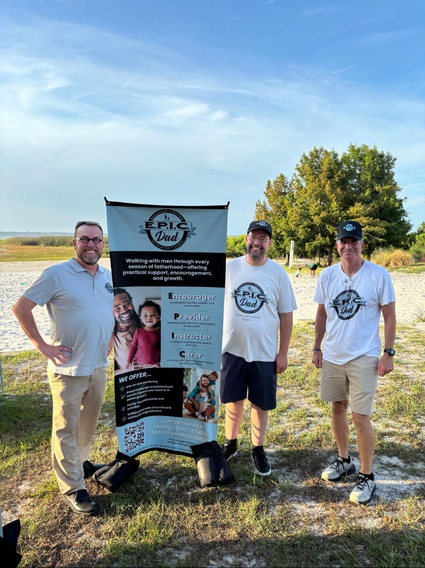 Three men stand in front of a banner with "EPIC DAD" printed on it, with a sandy beach and trees in the background under a clear sky.