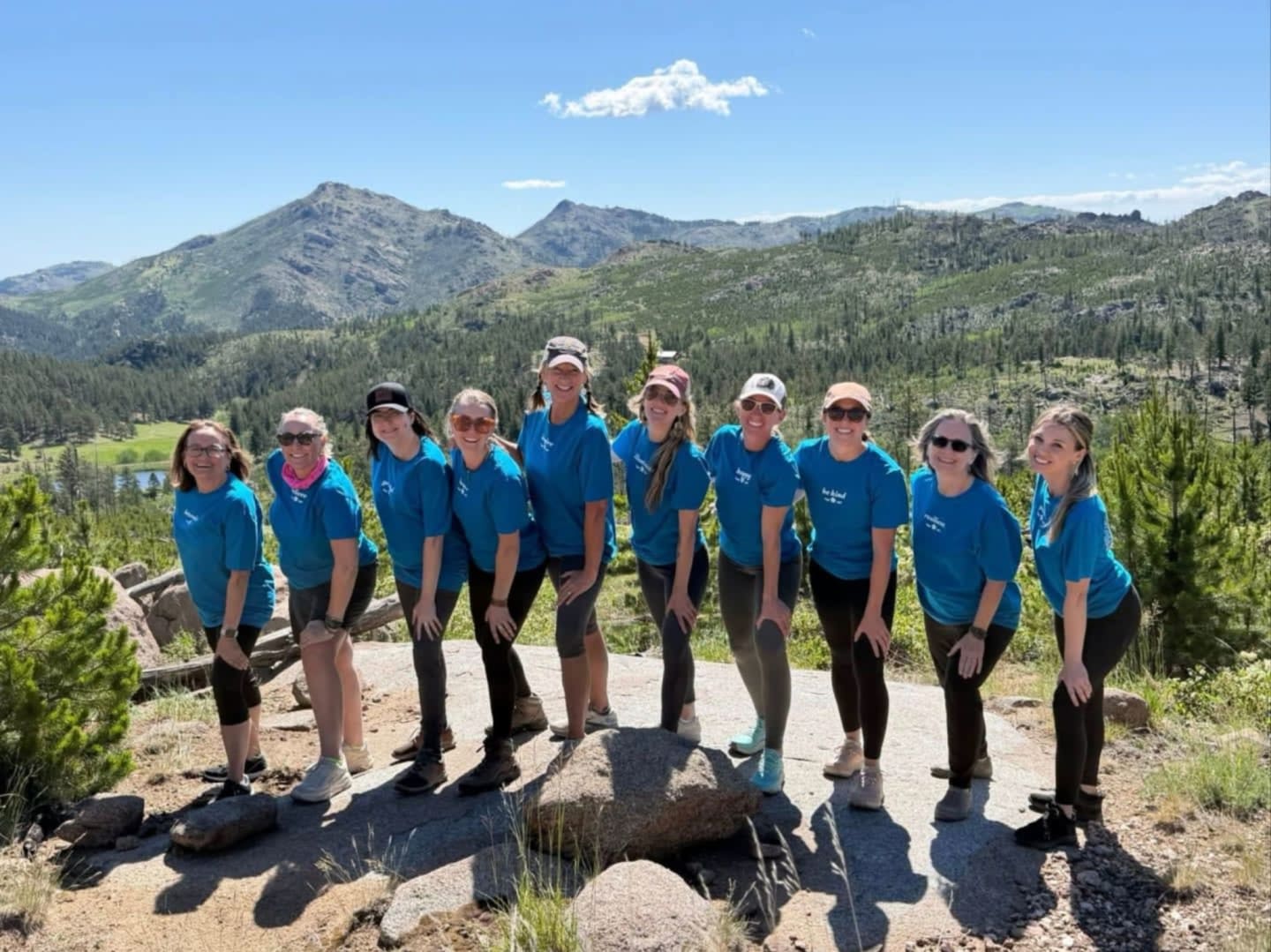 A group of nine women in matching blue shirts stand on a rocky outcrop in the foreground, with a vast, mountainous landscape dotted with pine trees and a distant lake stretching out behind them under a clear blue sky.