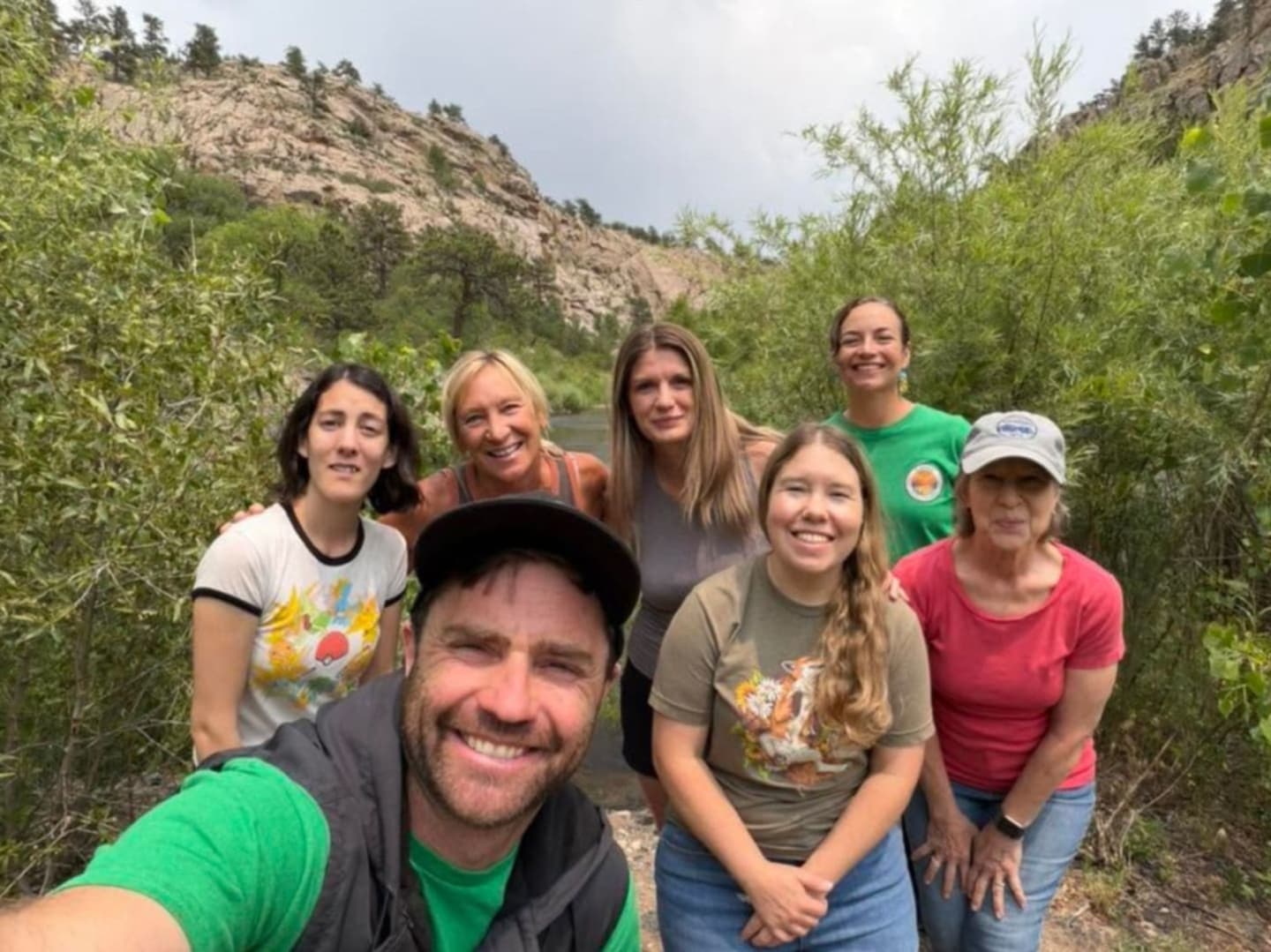 A group of six smiling people pose for a selfie in the foreground, with a river and rocky, tree-lined canyon in the background.
