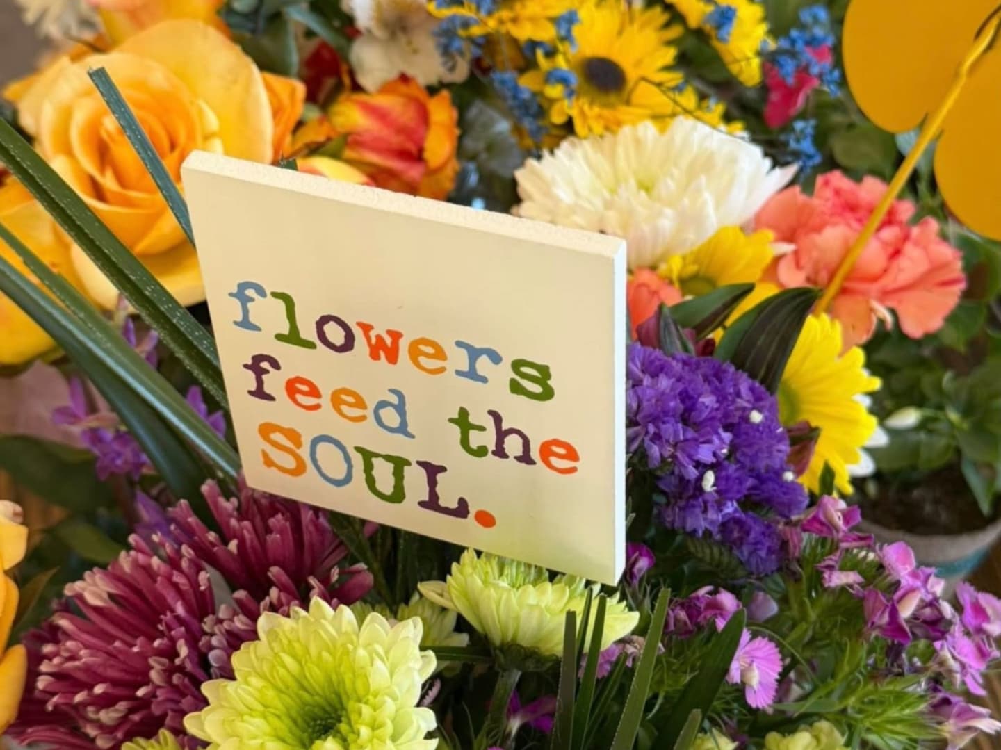 A vibrant bouquet of colorful flowers, featuring a sign that reads "flowers feed the soul," is set against a blurred background of more blooms and yellow butterfly decorations.