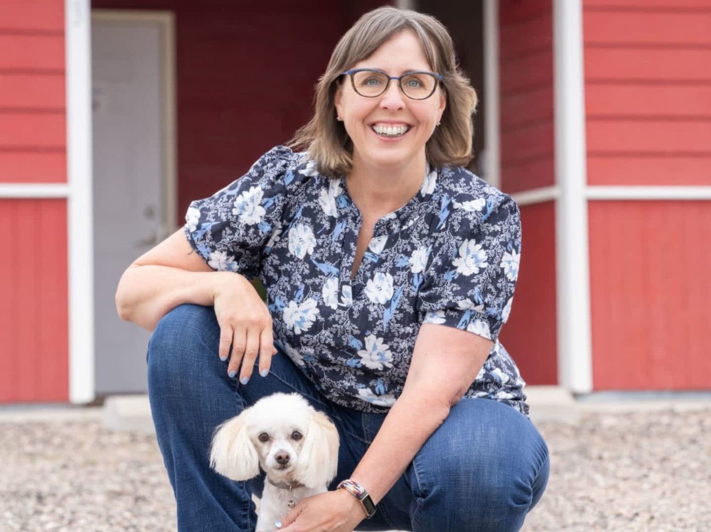 A woman in a floral shirt and jeans crouches beside a white poodle on a gravel path, with a red barn in the background.