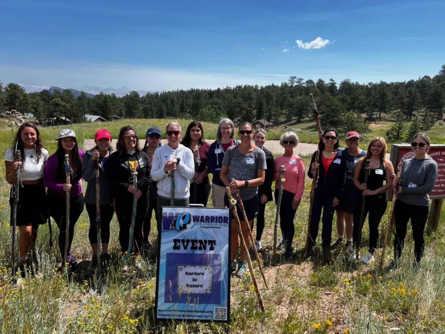 A group of women with hiking poles stand in a grassy field in front of a sign for a "Warrior" event, with a wooded hillside and blue sky in the background.