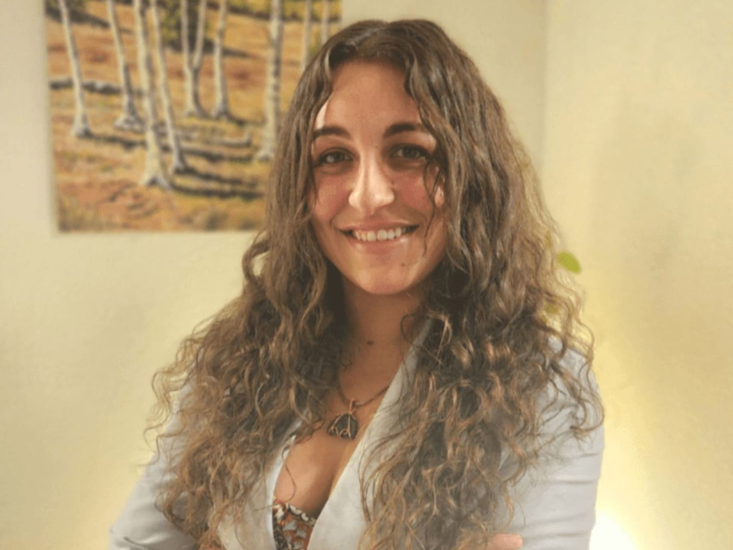 A smiling woman with long, curly brown hair and arms crossed in the foreground is set against a background featuring a painting of birch trees.