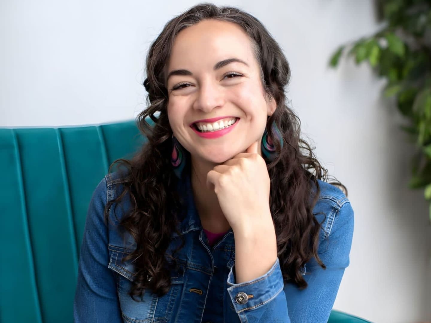 A smiling woman in a denim jacket rests her chin on her hand while sitting on a teal couch with a plant in the background.