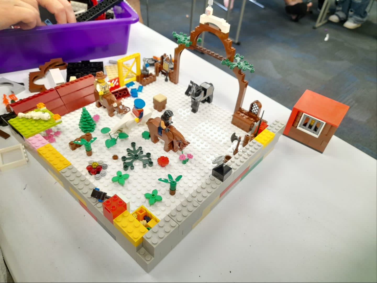 A LEGO diorama of a ranch scene with horses and cowboys in the foreground, and a purple bin of LEGO bricks and people's legs in the background.