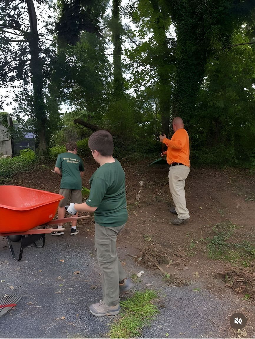 In the foreground, two boys in green shirts work with a wheelbarrow and rake, while in the background, a man in an orange shirt uses a weed trimmer among trees.