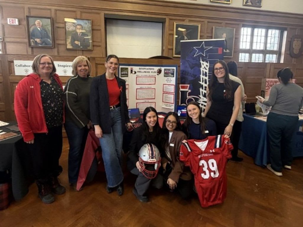 A group of women stand behind a table displaying a football helmet and jersey, with portraits and a large window visible in the wood-paneled background.