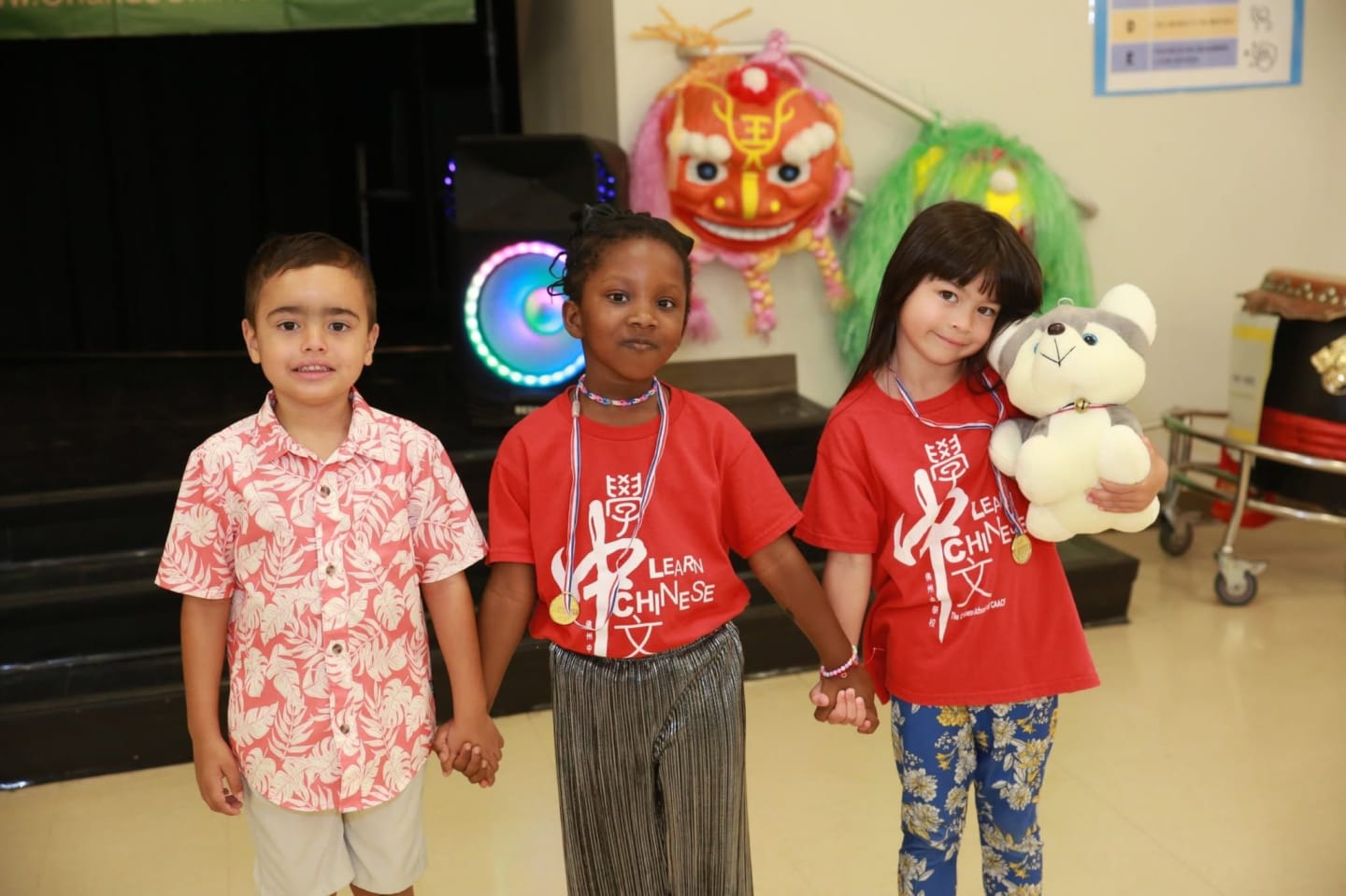 Three children, two girls and a boy, stand holding hands in the foreground, while colorful lion dance costumes and a speaker are visible in the background.