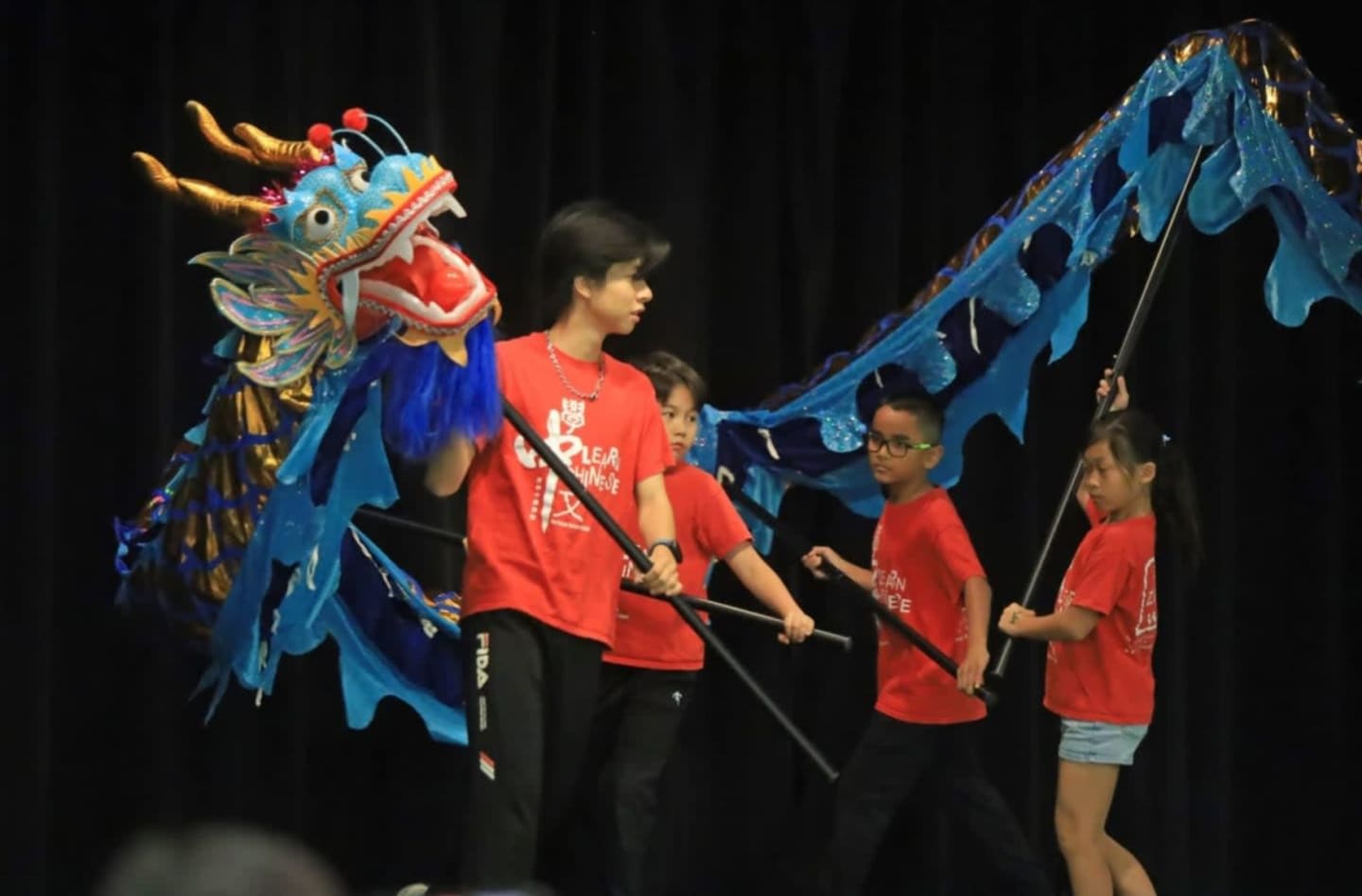 Children in red shirts carry a large blue dragon puppet across a dark stage.