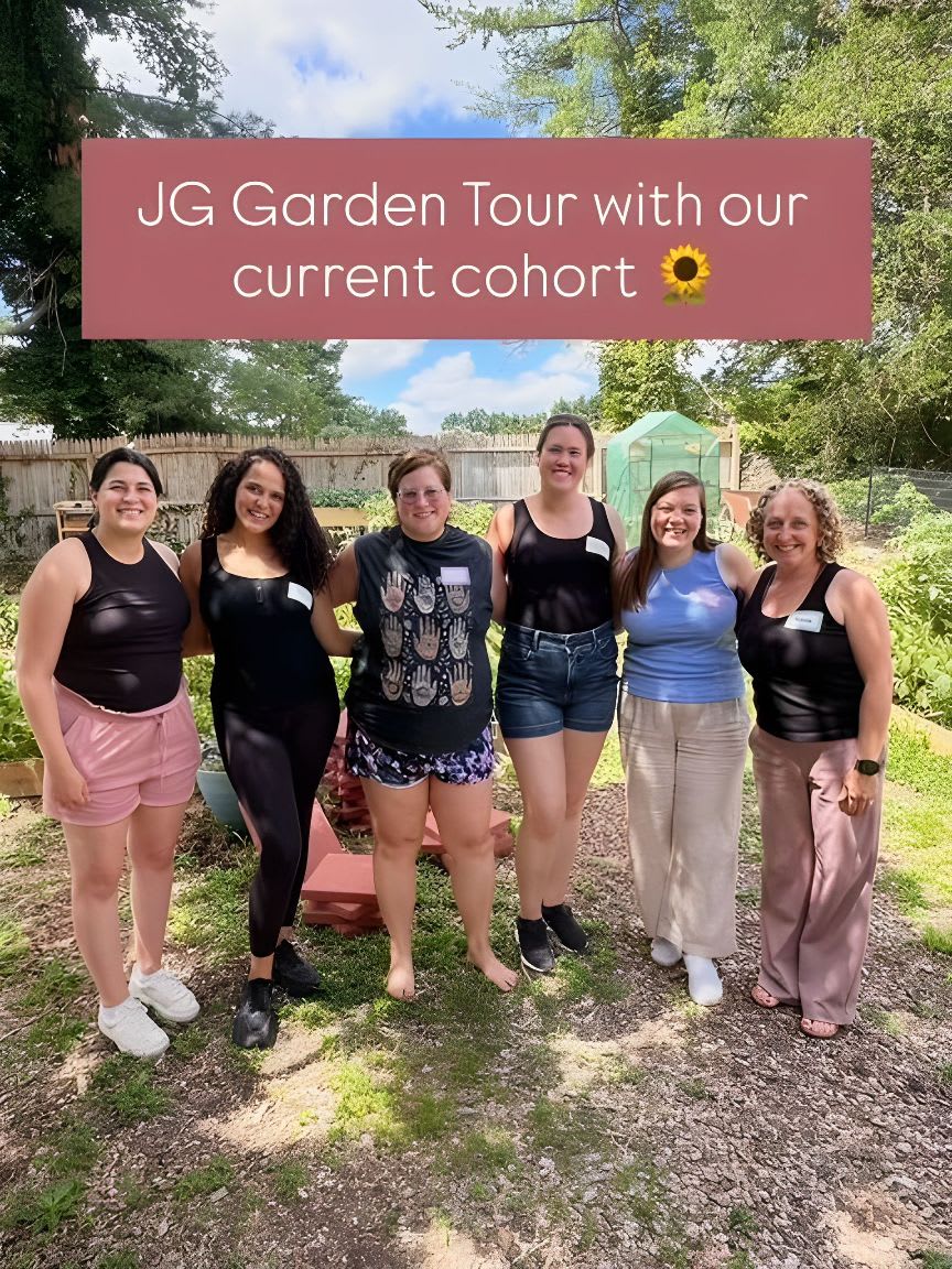 Six women stand in a garden with a wooden fence and greenery in the background.